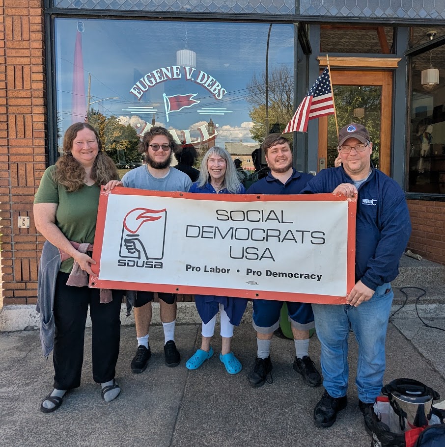 Group of five individuals holding a banner for Social Democrats USA outside a building with windows displaying a sign for Eugene V. Debs Hall.