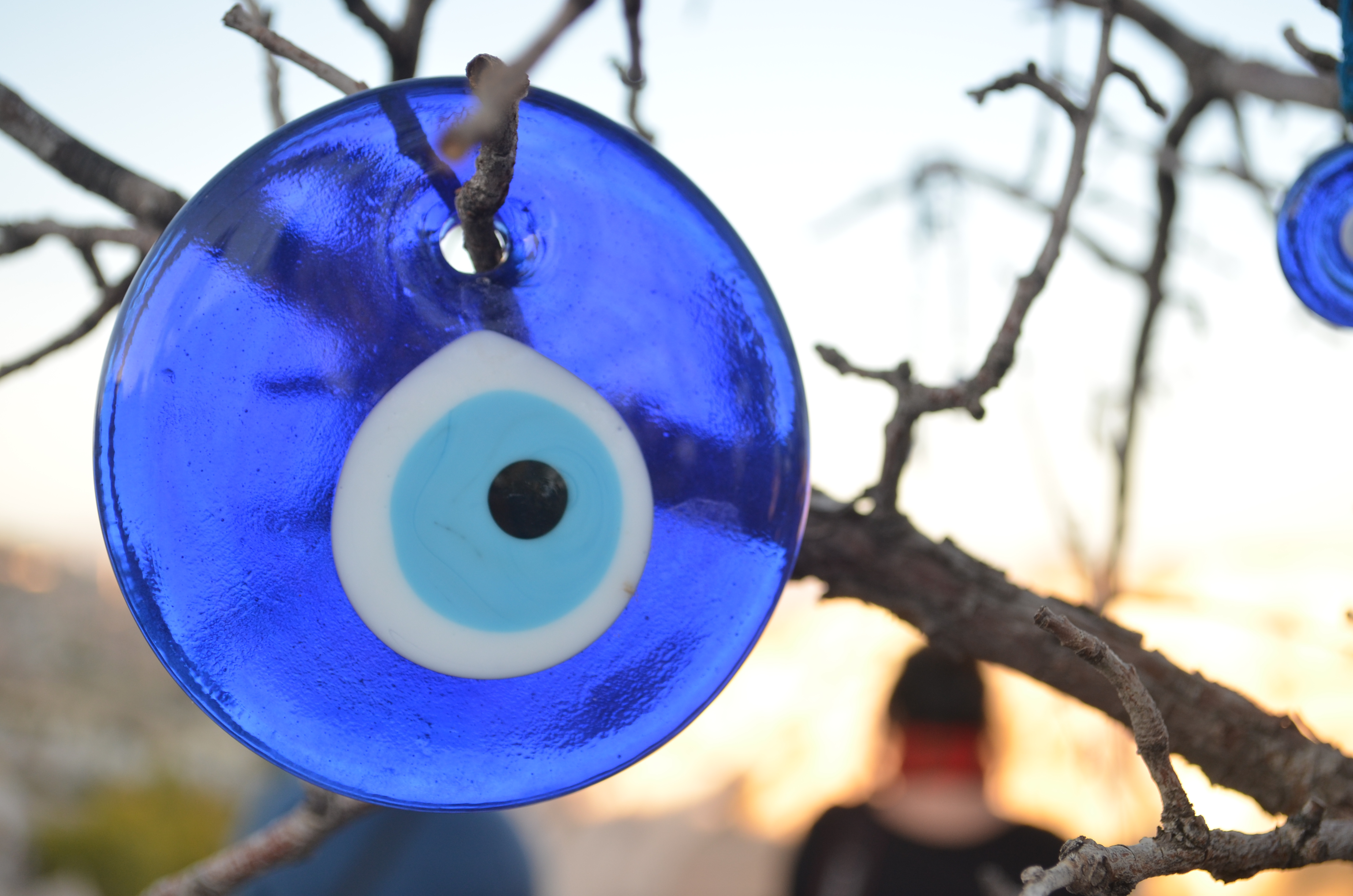 A close-up of a blue glass evil eye charm hanging from a branch, with a blurred background of a person and a sunset.