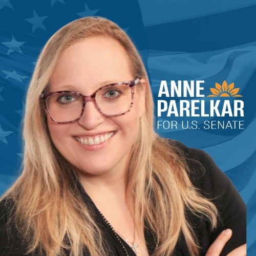 Portrait of a woman with long blonde hair and glasses smiling, with a blue background featuring an American flag and text that reads 'Anne Parelkar for U.S. Senate.'